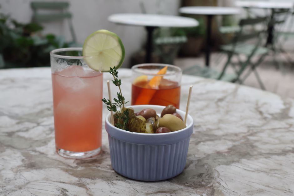 Two cocktails with lime and garnish beside a bowl of pickles on a marble table, outdoor setting in New York.