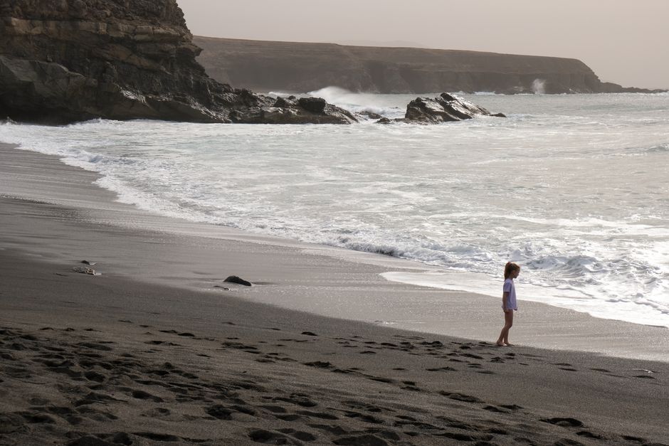A young girl stands at the edge of a rocky beach at dusk, gazing at the sea.