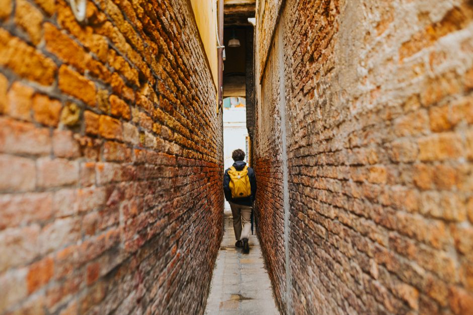 A person with a yellow backpack walks through a narrow alley between brick walls, capturing a sense of exploration.
