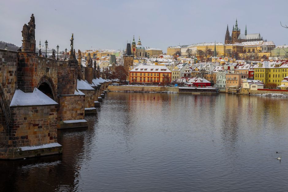 Snow-covered Charles Bridge and Prague Castle reflect in Vltava River on a winter day.