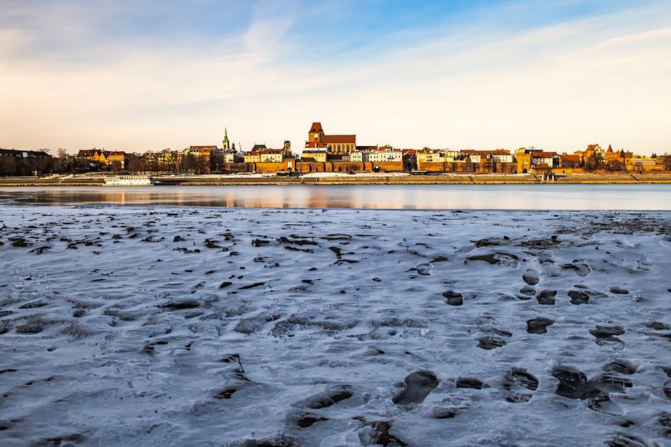 A snowy urban landscape with a historic city skyline reflecting on a tranquil river under a clear winter sky.