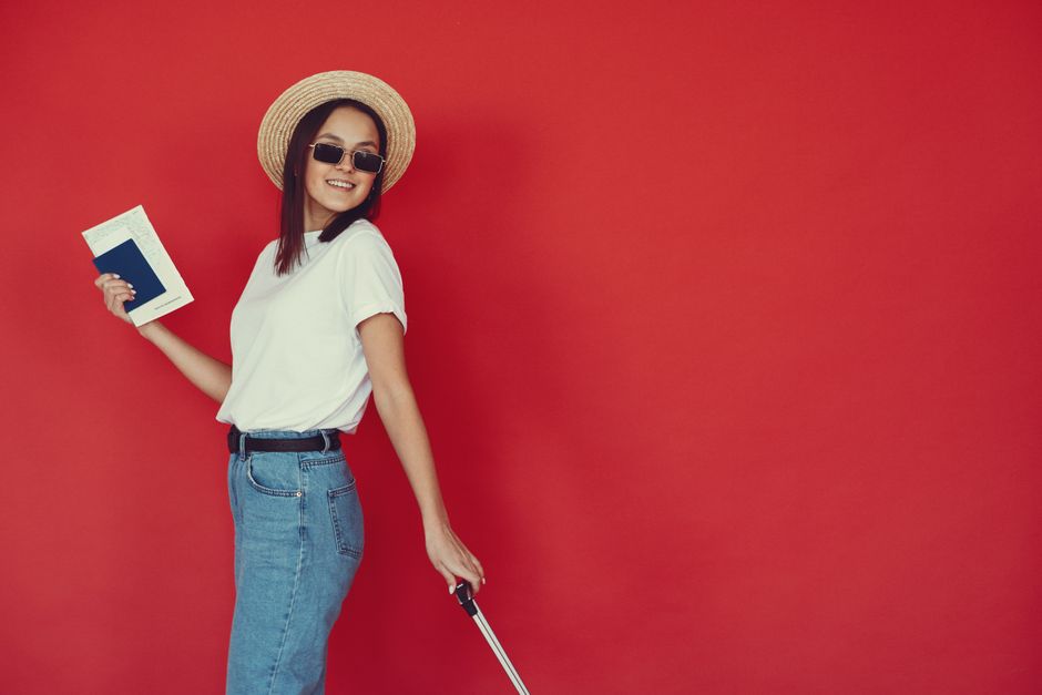 Cheerful young woman in stylish clothes and sunglasses smiling at camera and holding passport and tickets in hand on red background