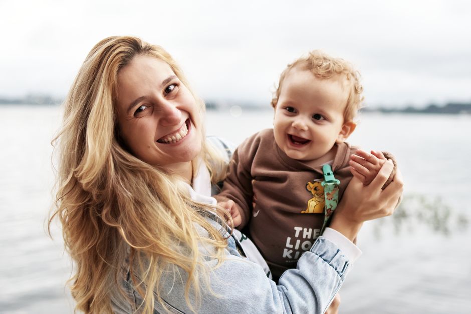A joyful mother holds her smiling baby by the waterfront on a cloudy day.
