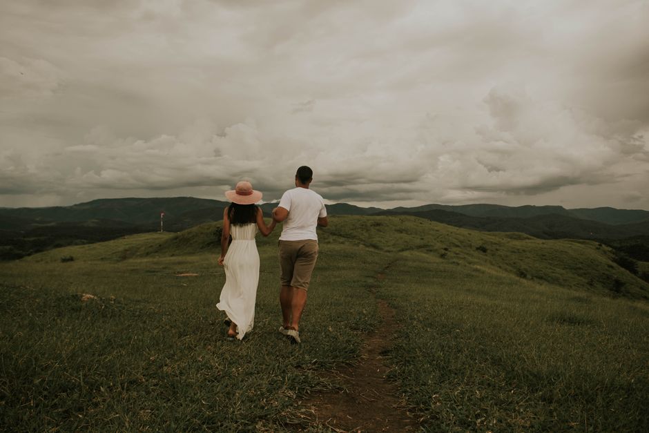 A couple walking hand in hand through a lush green hill under an overcast sky.