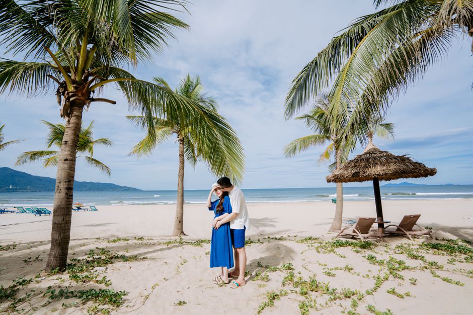 A couple shares a tender moment under palm trees on a sunny beach.