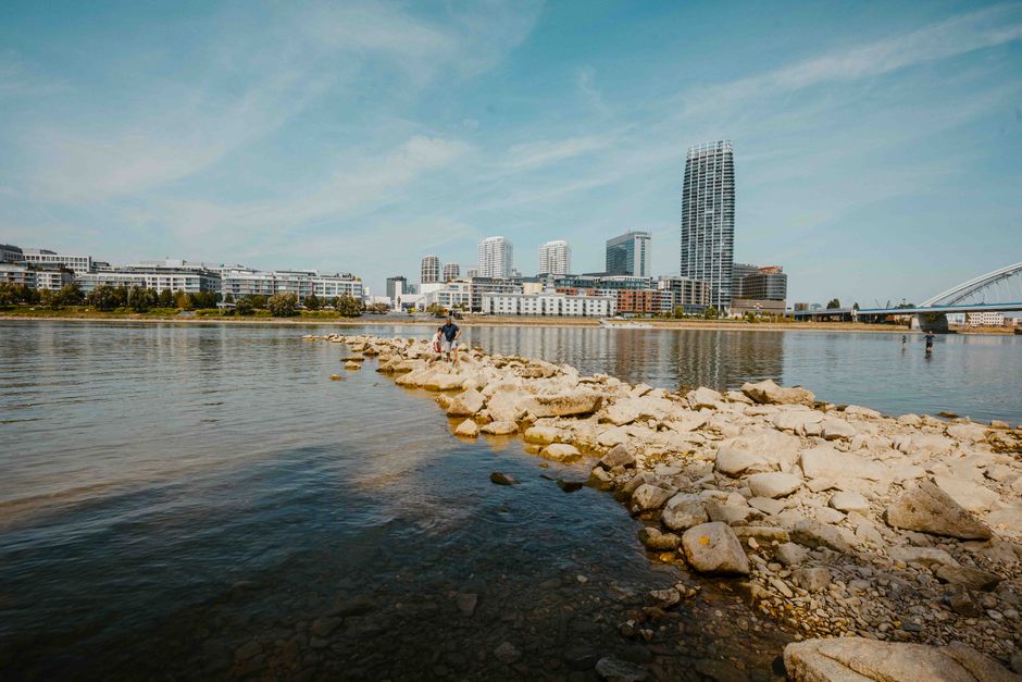 A clear view of a modern city skyline from a rocky riverside path with a bridge.