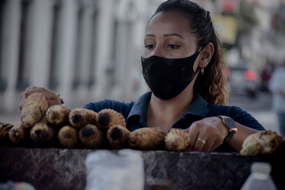 Woman vendor with face mask organizing grilled corn at a street stall during daytime.