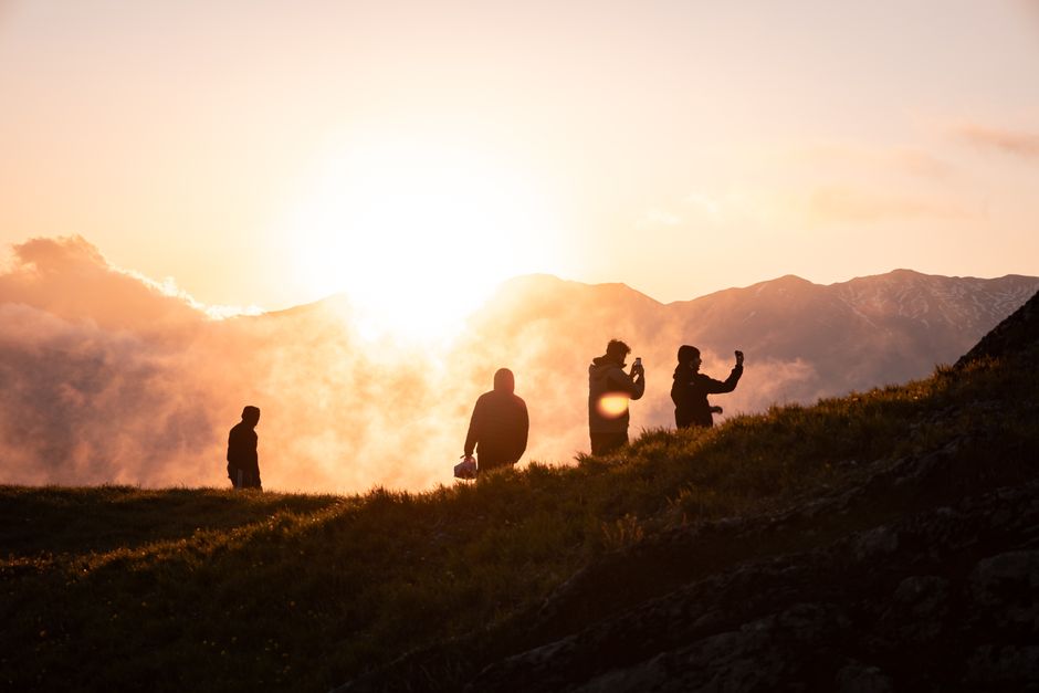 Four silhouetted hikers enjoy a breathtaking mountain sunset, capturing the moment with smartphones.