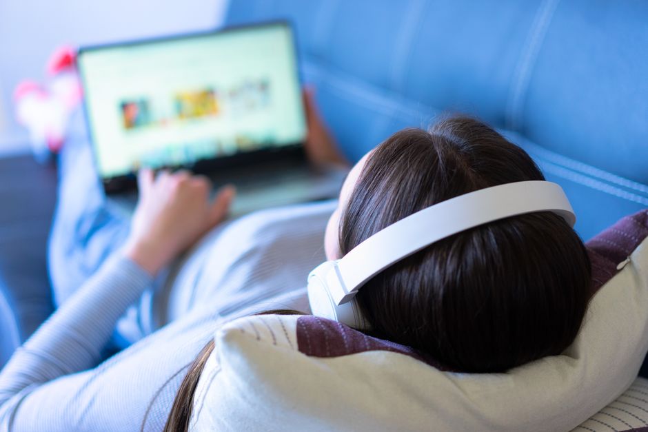 A woman lying on a sofa, enjoying music with headphones and browsing the internet on a laptop.