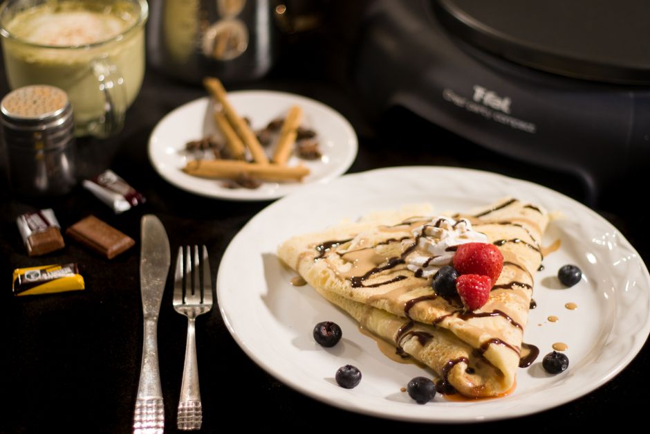 A tempting crepe topped with fresh blueberries, raspberries, and a chocolate drizzle on a white plate.