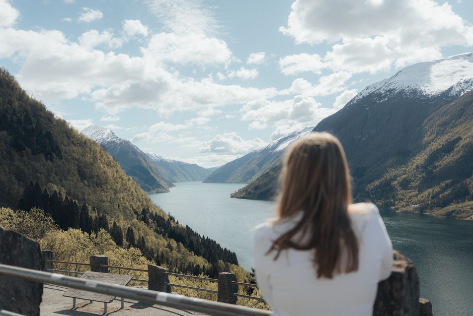 Woman enjoying a panoramic view of a Norwegian fjord surrounded by mountains, under a clear spring sky.