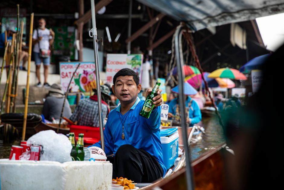 A man offers a beer bottle in a bustling Thailand floating market with colorful umbrellas and stalls.