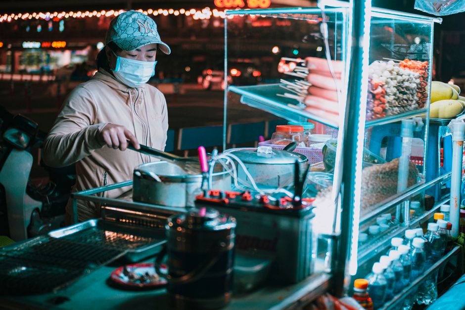 Street vendor in a mask preparing food at an illuminated night market stall.