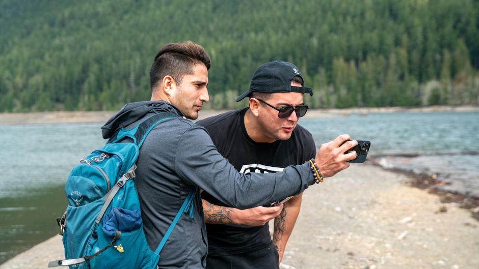 Two men enjoy a scenic outdoor adventure by the lake, capturing the moment with a smartphone.
